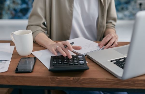 A person using a calculator beside a laptop and notepad