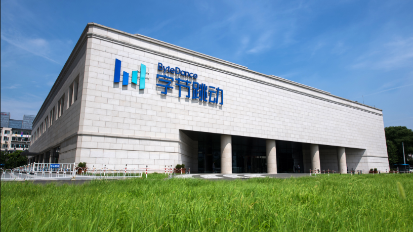 图片展示的是字节跳动的建筑外观。建筑主体为浅灰色,正面有“ByteDance”及“字节跳动”字样,左侧还有蓝色的“BD”标识。建筑前方是一片绿草地,背景天空晴朗,有几朵白云。图片与上下文的关系是,通过展示字节跳动的建筑,直观呈现了字节跳动这一科技企业形象,为理解其在AI产品中的角色提供背景信息。