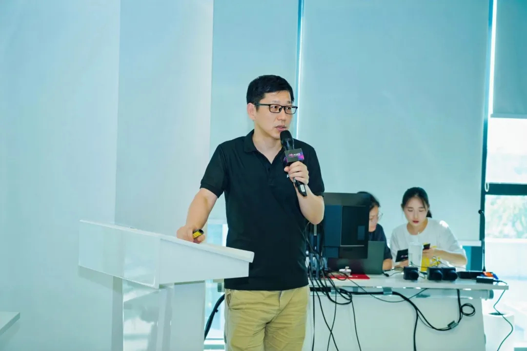 图片展示的是Pixmax技术总监赵志杰在Pixmax上海AIGC活动分享现场的场景。他身着黑色短袖,搭配浅色裤子,站在白色讲台旁,手持麦克风,专注地进行分享。讲台后方有电脑、耳机等设备,旁边还有两位工作人员,一位在操作电脑,另一位在整理物品。背景为明亮的室内环境,窗外有绿植。此图与上下文紧密相关,直观呈现了赵志杰分享时的现场情况。