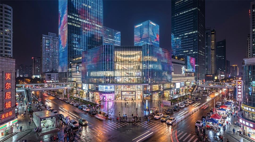 图片展示的是现代商业综合体夜景,建筑以玻璃幕墙为主,灯光效果绚丽。画面中有多栋高楼大厦,其中一栋建筑顶部有色彩斑斓的灯光装饰。街道上车流不息,路边有行人,远处可见其他建筑和城市景观。该图片是示例1中“现代商业综合体,玻璃幕墙建筑,夜景灯光,城市街道环境,高质量写实建筑摄影风格,广角镜头”提示词生成效果的展示,直观呈现了AI模型生成“PixNano Pro”生成的建筑效果图。